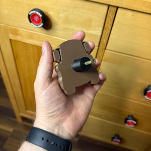 May include: A brown cabinet knob in the shape of a car. The knob is being held up in front of a wooden cabinet. Other cabinet knobs are visible, including one with a red and black design.