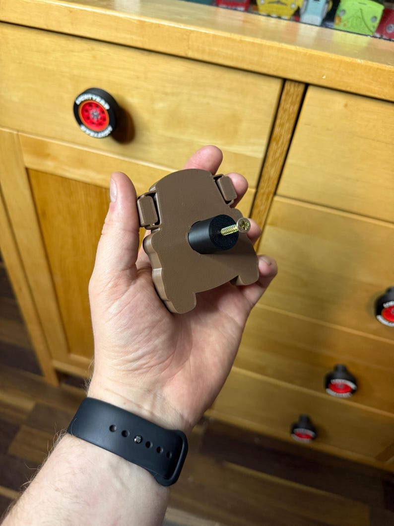 May include: A brown cabinet knob in the shape of a car. The knob is being held up in front of a wooden cabinet. Other cabinet knobs are visible, including one with a red and black design.