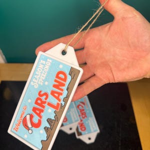 K&ouml;nnte beinhalten: Rechteckiges wei&szlig;es Etikett mit dem Text "Welcome to Cars Land" in Rot und Braun, mit einem Weihnachtsmannhut. Das Etikett hat einen hellblauen Hintergrund mit dem Text "Season's Speedings". Ein brauner Faden ist am Etikett befestigt.