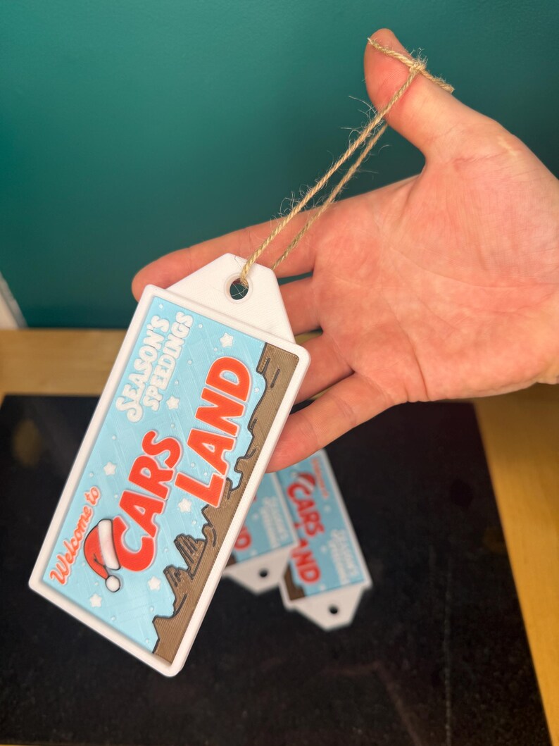 K&ouml;nnte beinhalten: Rechteckiges wei&szlig;es Etikett mit dem Text "Welcome to Cars Land" in Rot und Braun, mit einem Weihnachtsmannhut. Das Etikett hat einen hellblauen Hintergrund mit dem Text "Season's Speedings". Ein brauner Faden ist am Etikett befestigt.