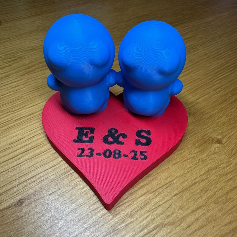 Cute 3D-printed wedding cake topper featuring two blue figurines on a red heart base engraved with initials and a date. Ideal for weddings, anniversaries, or events, this cute eco-friendly PLA decoration is a unique keepsake for special occasions.
