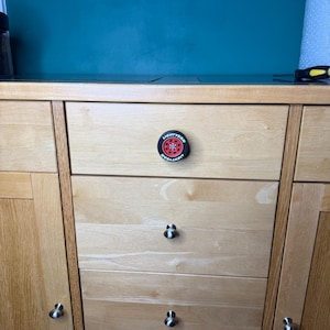 May include: A light wood cabinet with multiple drawers and doors. The center drawer features a round, black and red knob with the text "LIGHTYEAR" around the edge and a red wheel design in the center. Other drawers have silver knobs.