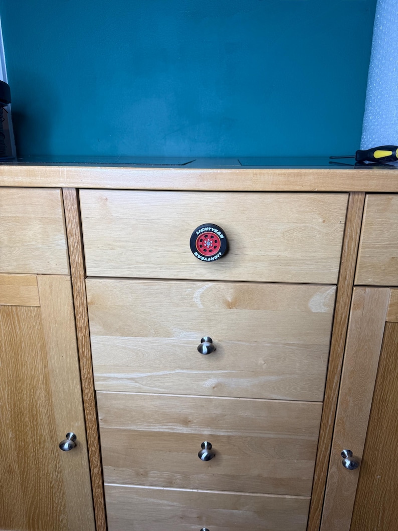 May include: A light wood cabinet with multiple drawers and doors. The center drawer features a round, black and red knob with the text "LIGHTYEAR" around the edge and a red wheel design in the center. Other drawers have silver knobs.