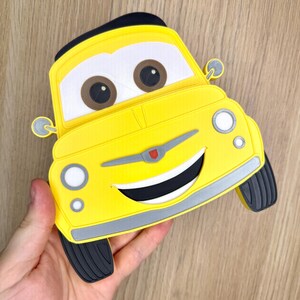 May include: A yellow car-shaped accessory with a smiling face, brown eyes, and black tires. The car has a black top and a silver trim. The design is reminiscent of a cartoon character.