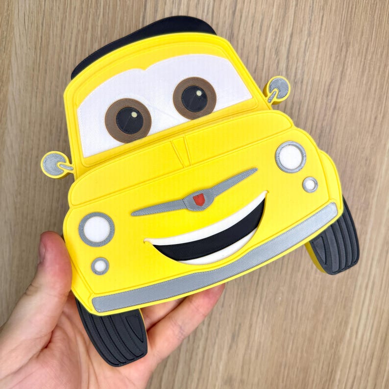 May include: A yellow car-shaped accessory with a smiling face, brown eyes, and black tires. The car has a black top and a silver trim. The design is reminiscent of a cartoon character.