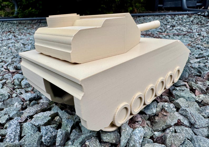 May include: A beige 3D printed model of a tank featuring a turret, a long barrel, and a series of circular wheels along the side. The tank has a matte finish with visible horizontal layer lines from the printing process and a geometric structure.