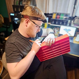 May include: A person wearing a headlamp and glasses is working on a red plastic organizer with multiple compartments. The person is holding a pen and appears to be marking the organizer. The background includes a 3D printer and spools of filament.
