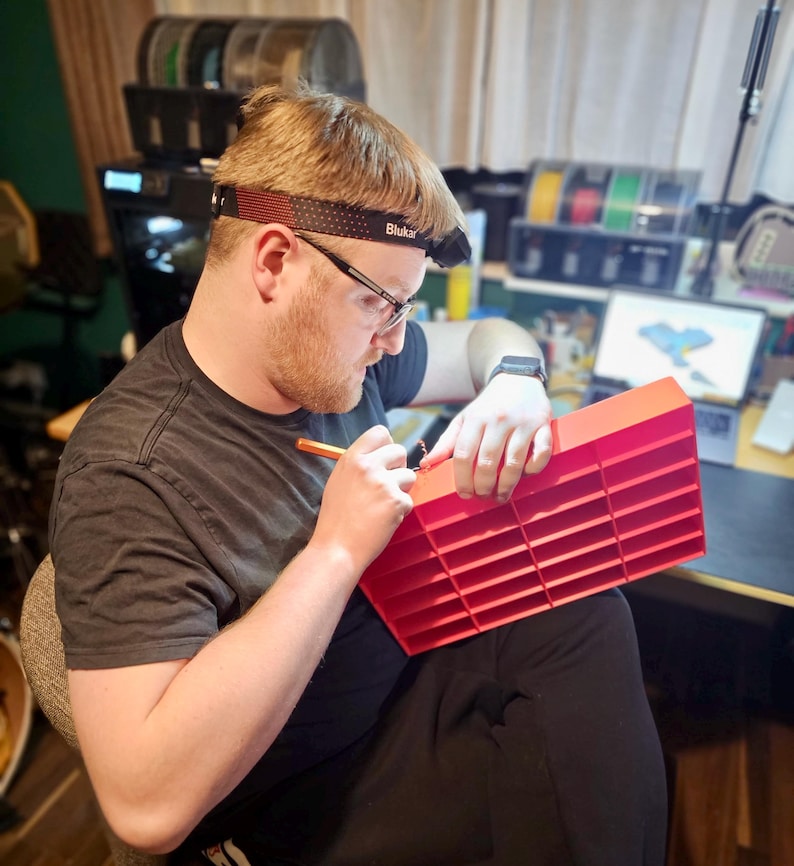May include: A person wearing a headlamp and glasses is working on a red plastic organizer with multiple compartments. The person is holding a pen and appears to be marking the organizer. The background includes a 3D printer and spools of filament.