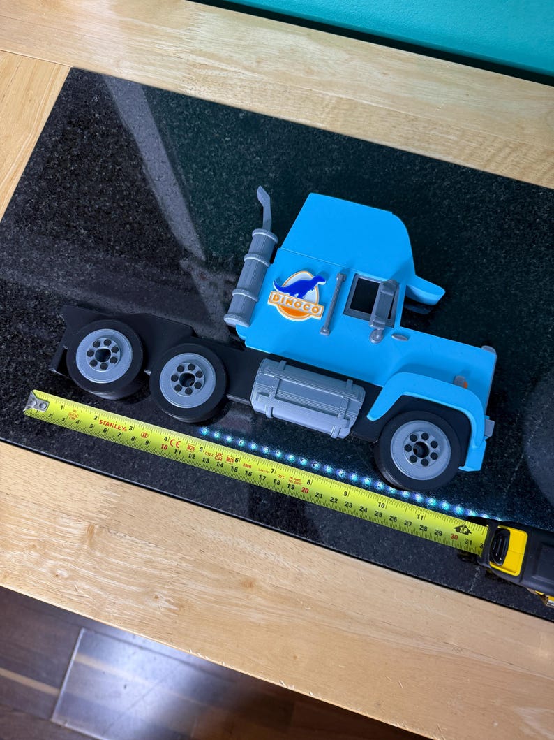 May include: A light blue toy semi-truck with black wheels and a silver exhaust stack. The truck has the word "DINOCO" printed on the side. A yellow tape measure is next to the truck.
