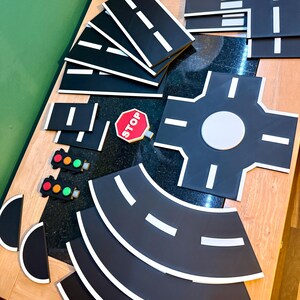 May include: A collection of black road pieces with white lane markings, a red stop sign, and traffic lights. The set includes straight, curved, and intersection pieces for creating a toy road system. The pieces are arranged on a wooden surface.
