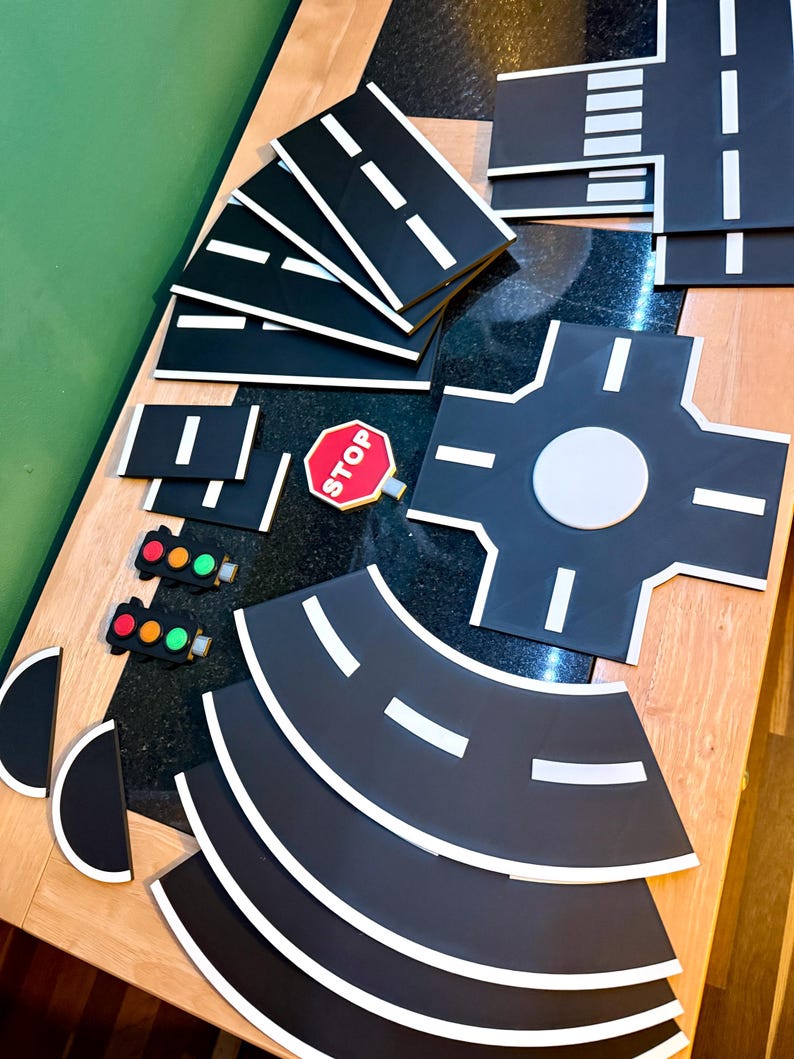 May include: A collection of black road pieces with white lane markings, a red stop sign, and traffic lights. The set includes straight, curved, and intersection pieces for creating a toy road system. The pieces are arranged on a wooden surface.