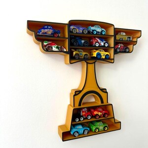 Puede incluir: Estanter&iacute;a de pared amarilla con forma de trofeo con m&uacute;ltiples compartimentos, cada uno con una colecci&oacute;n de coches de carreras de juguete. La estanter&iacute;a tiene un borde negro y est&aacute; montada en una pared blanca. Los coches son de colores brillantes.