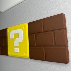 Op de afbeelding: Een wanddecoratie met elementen uit de Super Mario-videogameserie. Inclusief een gele vraagtekenblok met een wit vraagteken, bruine bakstenen en een witte wolk. De items zijn 3D-geprint en op een grijze muur gemonteerd.