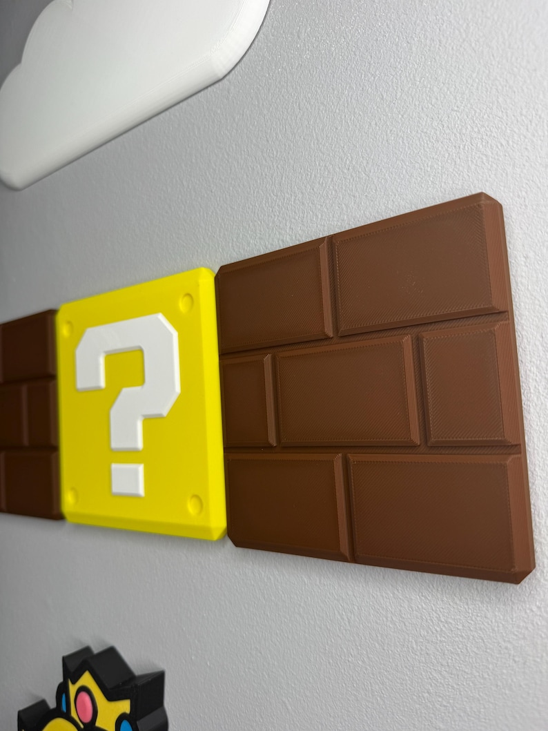 Op de afbeelding: Een wanddecoratie met elementen uit de Super Mario-videogameserie. Inclusief een gele vraagtekenblok met een wit vraagteken, bruine bakstenen en een witte wolk. De items zijn 3D-geprint en op een grijze muur gemonteerd.