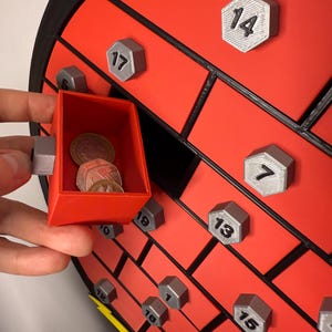 May include: A red and black advent calendar with a brick design. The calendar has numbered silver hexagonal doors. A small red drawer is open, revealing coins inside. The calendar is a festive item.