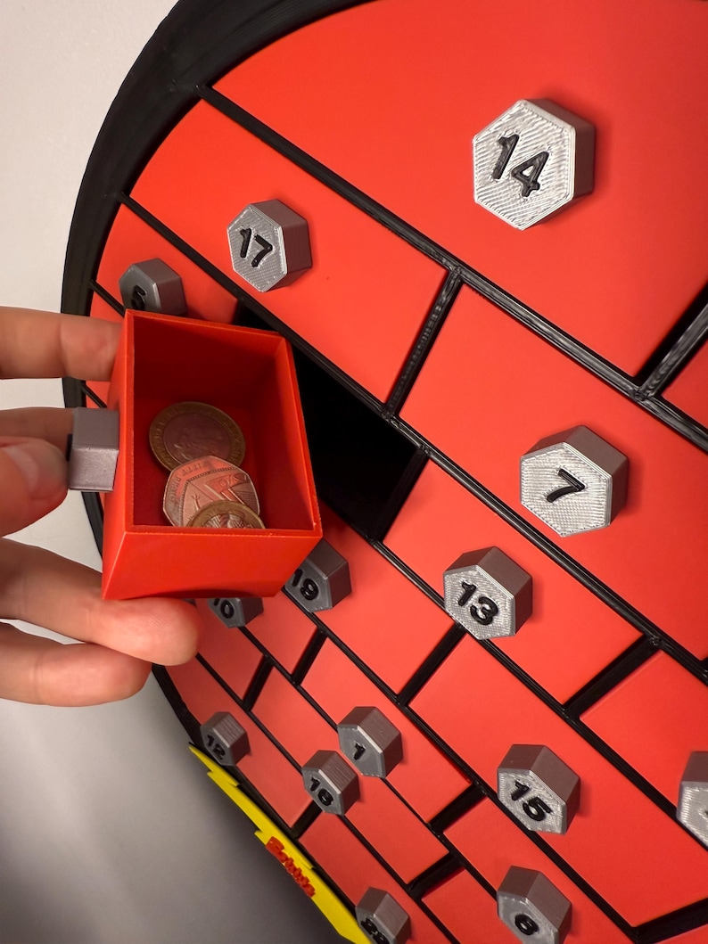 May include: A red and black advent calendar with a brick design. The calendar has numbered silver hexagonal doors. A small red drawer is open, revealing coins inside. The calendar is a festive item.