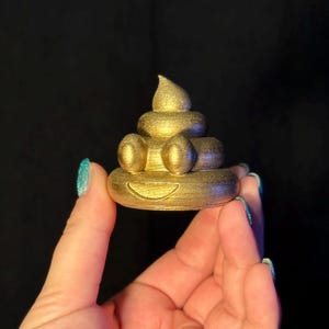 A hand with glittery blue nails holds a 5cm tall 3D-printed poop emoji figurine, painted gold and covered in fine glitter. The swirled design has raised eyes and a smiling mouth. Black background for contrast.