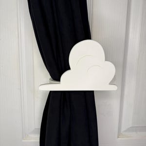 May include: A white cloud-shaped curtain tieback holds a black curtain against a white door. The tieback is made of a solid material and has a simple, clean design. The curtain is made of a dark fabric.