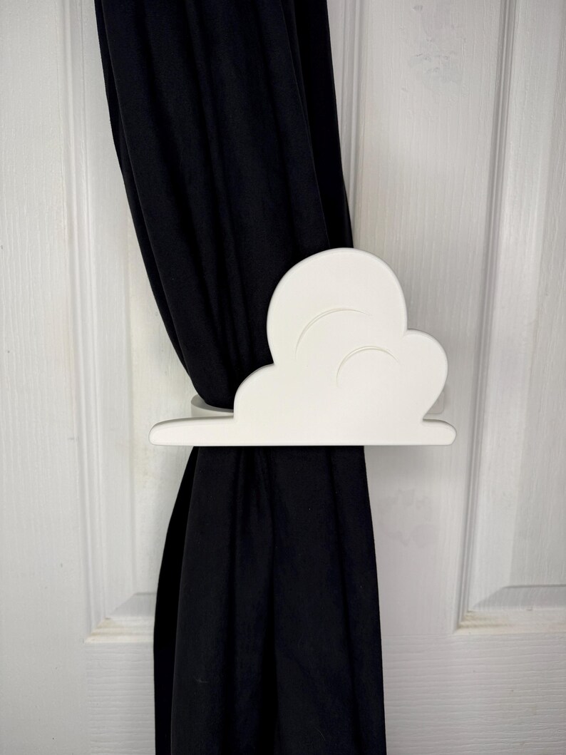 May include: A white cloud-shaped curtain tieback holds a black curtain against a white door. The tieback is made of a solid material and has a simple, clean design. The curtain is made of a dark fabric.