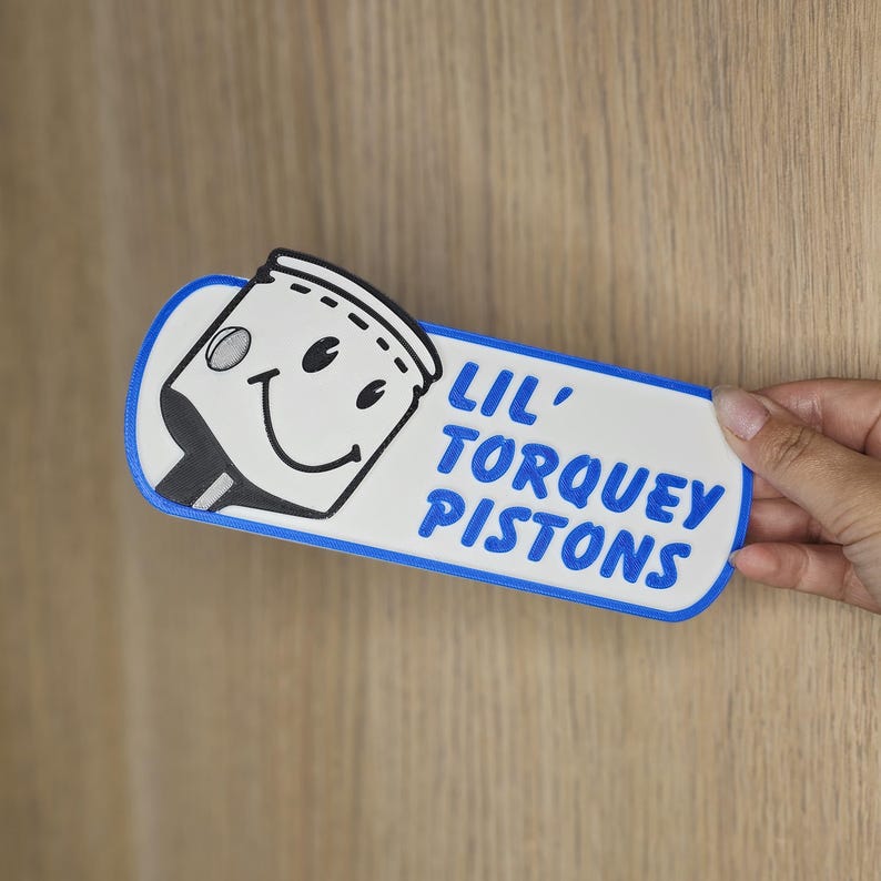 May include: A white and blue sign with the text "LIL' TORQUEY PISTONS" and a cartoon piston character. The sign has a blue border and is held by a hand.