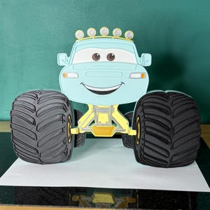Op de afbeelding: Een lichtblauwe monstertruck met grote zwarte banden en gele ophanging. De truck heeft een lachend gezicht, bruine ogen en een rij gele lampen. Het voertuig is gemaakt van papier of een vergelijkbaar materiaal.