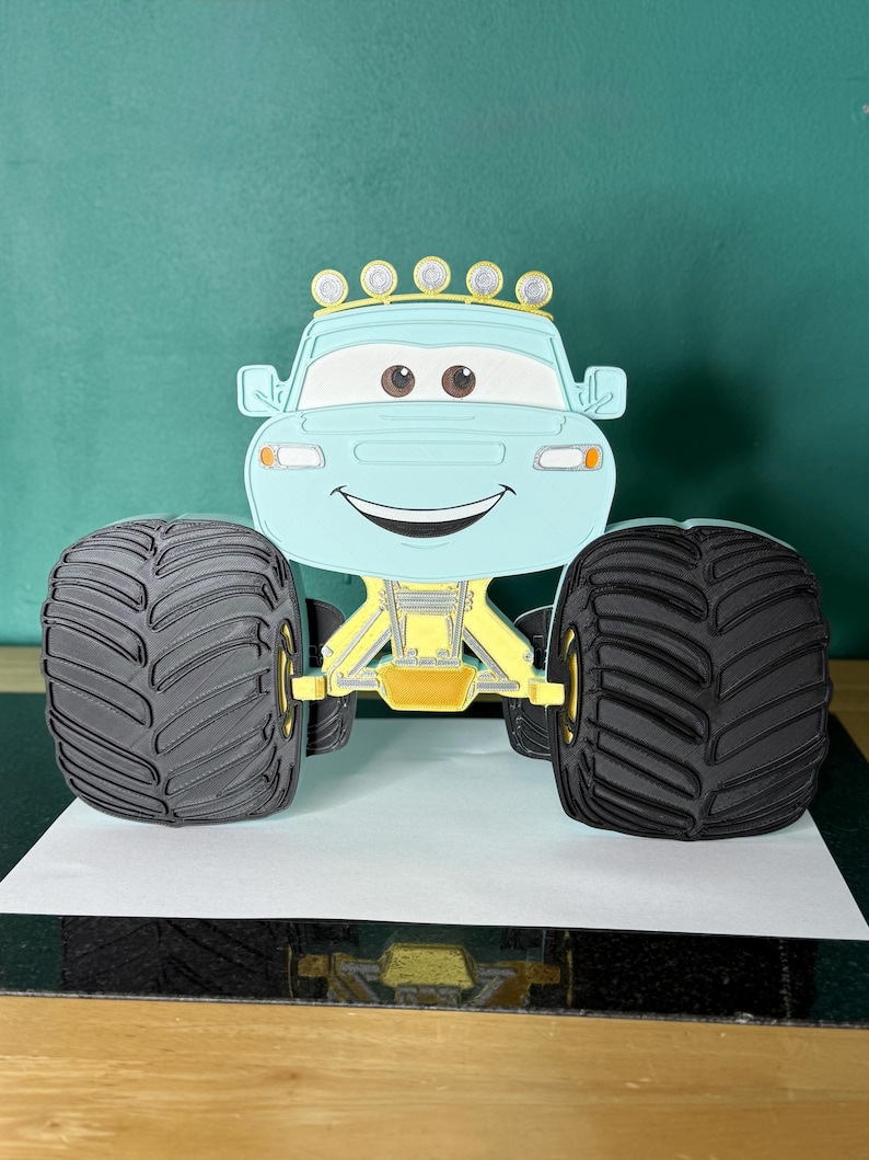Op de afbeelding: Een lichtblauwe monstertruck met grote zwarte banden en gele ophanging. De truck heeft een lachend gezicht, bruine ogen en een rij gele lampen. Het voertuig is gemaakt van papier of een vergelijkbaar materiaal.