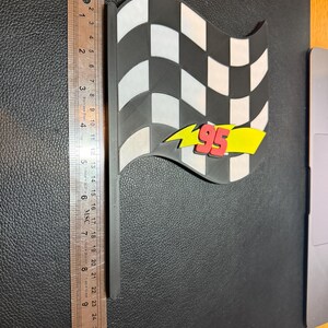 May include: A black and white checkered flag with a black handle. The flag has a yellow lightning bolt with the number 95 in red. The flag measures approximately 10 inches in length.