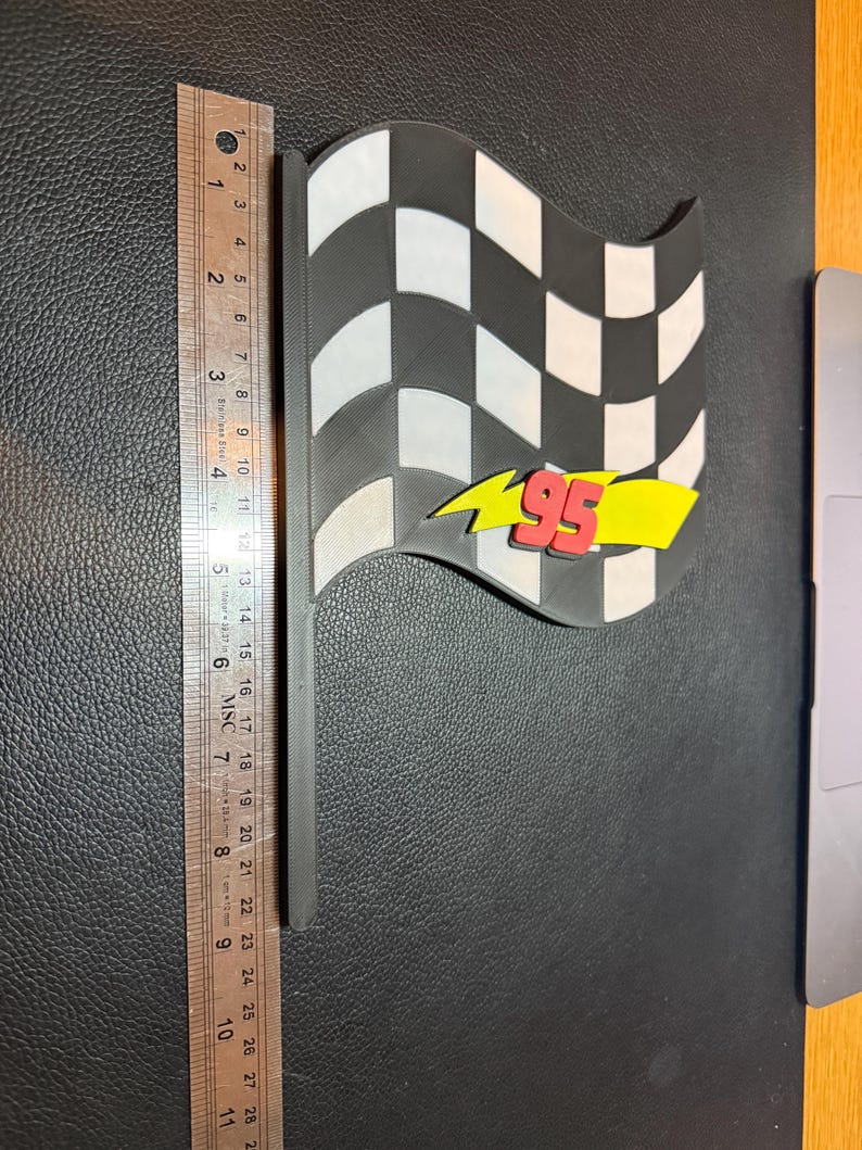 May include: A black and white checkered flag with a black handle. The flag has a yellow lightning bolt with the number 95 in red. The flag measures approximately 10 inches in length.