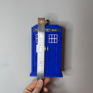 May include: A blue miniature police box with the text "PUBLIC CALL BOX" on top. The model is approximately 8 inches tall, with a ruler held up against it. The box has white window details and a small lamp on top.