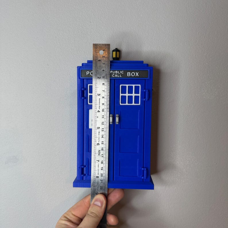 May include: A blue miniature police box with the text "PUBLIC CALL BOX" on top. The model is approximately 8 inches tall, with a ruler held up against it. The box has white window details and a small lamp on top.
