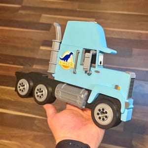 May include: A light blue toy truck with the Dinoco logo, held in a hand. The truck has black wheels, a silver exhaust pipe, and a silver fuel tank. The truck is approximately 12 inches long.