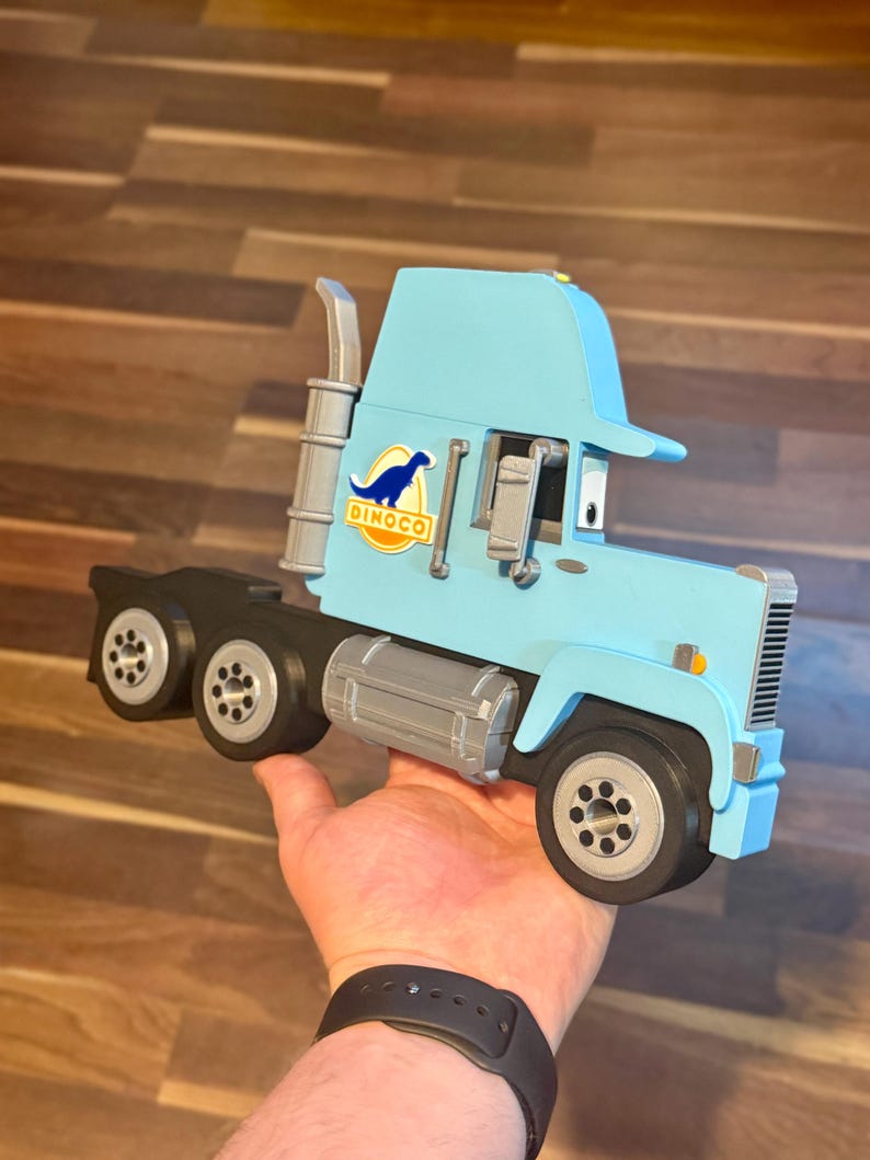 May include: A light blue toy truck with the Dinoco logo, held in a hand. The truck has black wheels, a silver exhaust pipe, and a silver fuel tank. The truck is approximately 12 inches long.