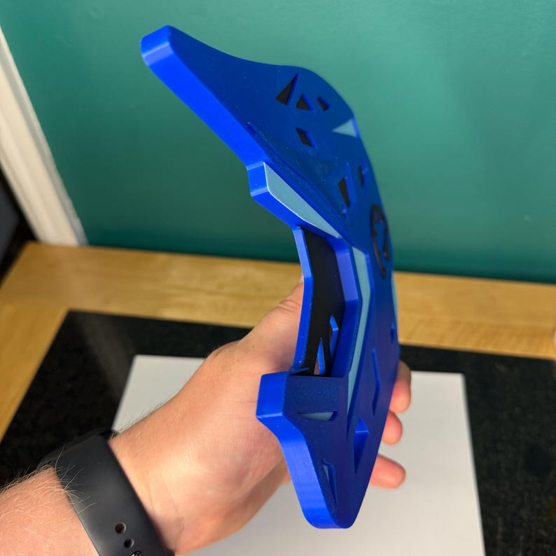 May include: A blue, geometric-shaped object with a curved design. The object has a series of cut-out shapes and a lighter blue accent. The object is held in a hand, and the background is a solid green color.