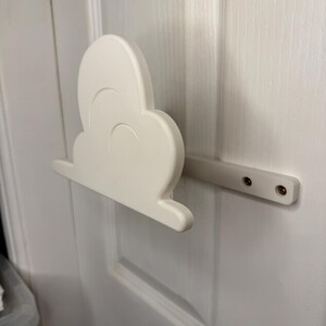 May include: A white cloud-shaped door pull, mounted on a white door. The cloud has a rounded shape with curved details and is attached to a rectangular bar with screws. The door is painted white.