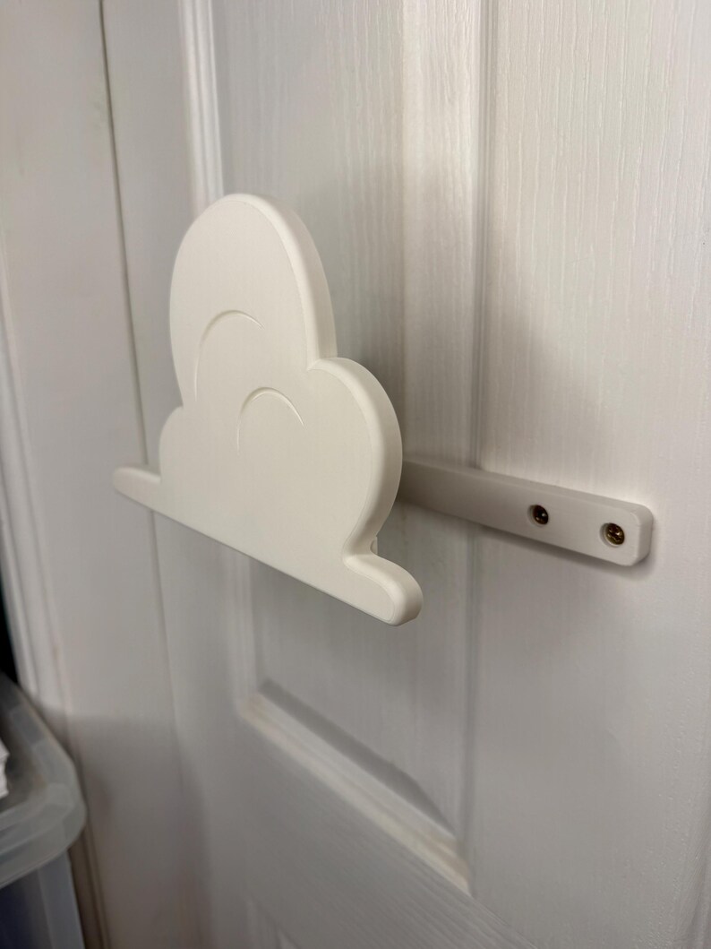 May include: A white cloud-shaped door pull, mounted on a white door. The cloud has a rounded shape with curved details and is attached to a rectangular bar with screws. The door is painted white.
