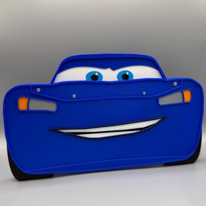 May include: A blue Lightning McQueen-themed toy car with a smiling face, white eyes, and blue irises. The car has black tires and orange and silver accents. The toy is designed to resemble the character from the animated movie "Cars".