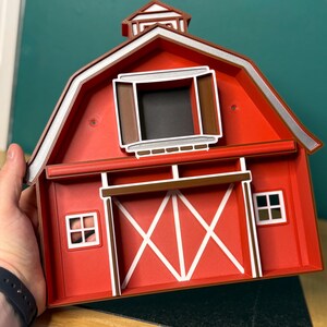Puede incluir: Una caja decorativa roja en forma de granero con detalles en blanco y marr&oacute;n. El granero presenta un borde de techo blanco, una ventana marr&oacute;n y un dise&ntilde;o de puerta blanco. Dos peque&ntilde;as ventanas con marco blanco est&aacute;n en los lados. La caja est&aacute; sostenida por una persona.