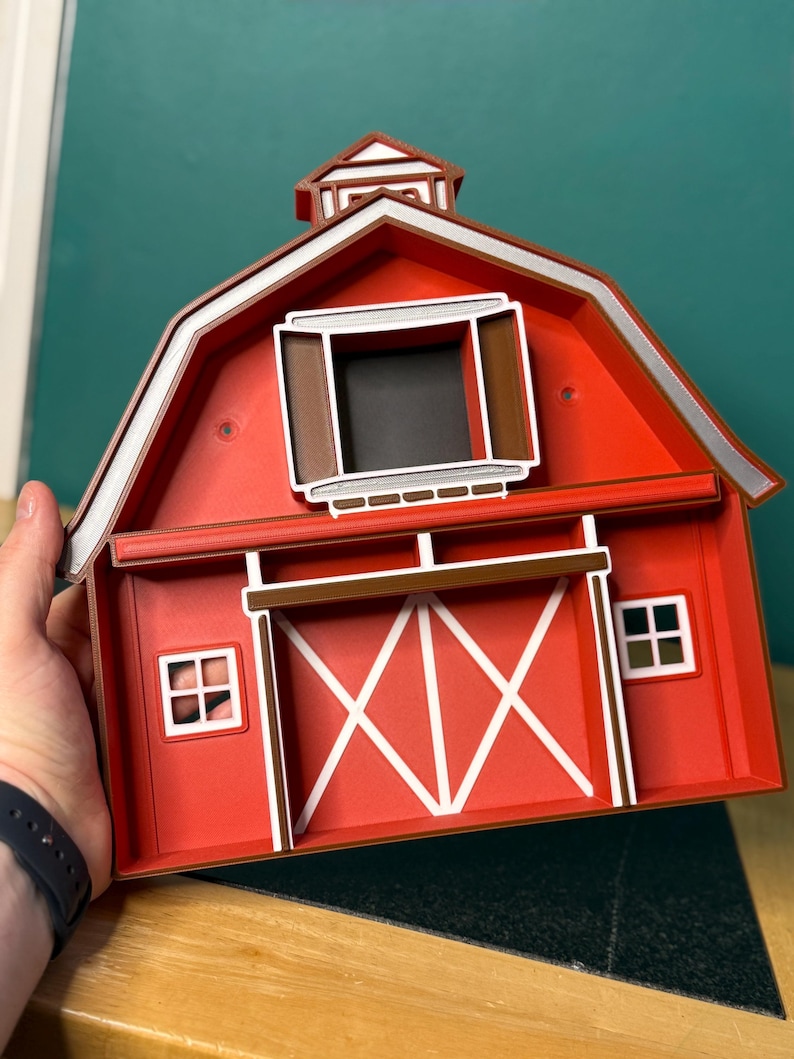 Puede incluir: Una caja decorativa roja en forma de granero con detalles en blanco y marr&oacute;n. El granero presenta un borde de techo blanco, una ventana marr&oacute;n y un dise&ntilde;o de puerta blanco. Dos peque&ntilde;as ventanas con marco blanco est&aacute;n en los lados. La caja est&aacute; sostenida por una persona.