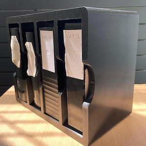 May include: A black, multi-compartment organizer with four sections. Each section holds a dark-colored bottle or item, with white labels reading "CAT 7" and "CAT 1". The organizer is made of a solid, dark material and sits on a light-colored wooden surface.