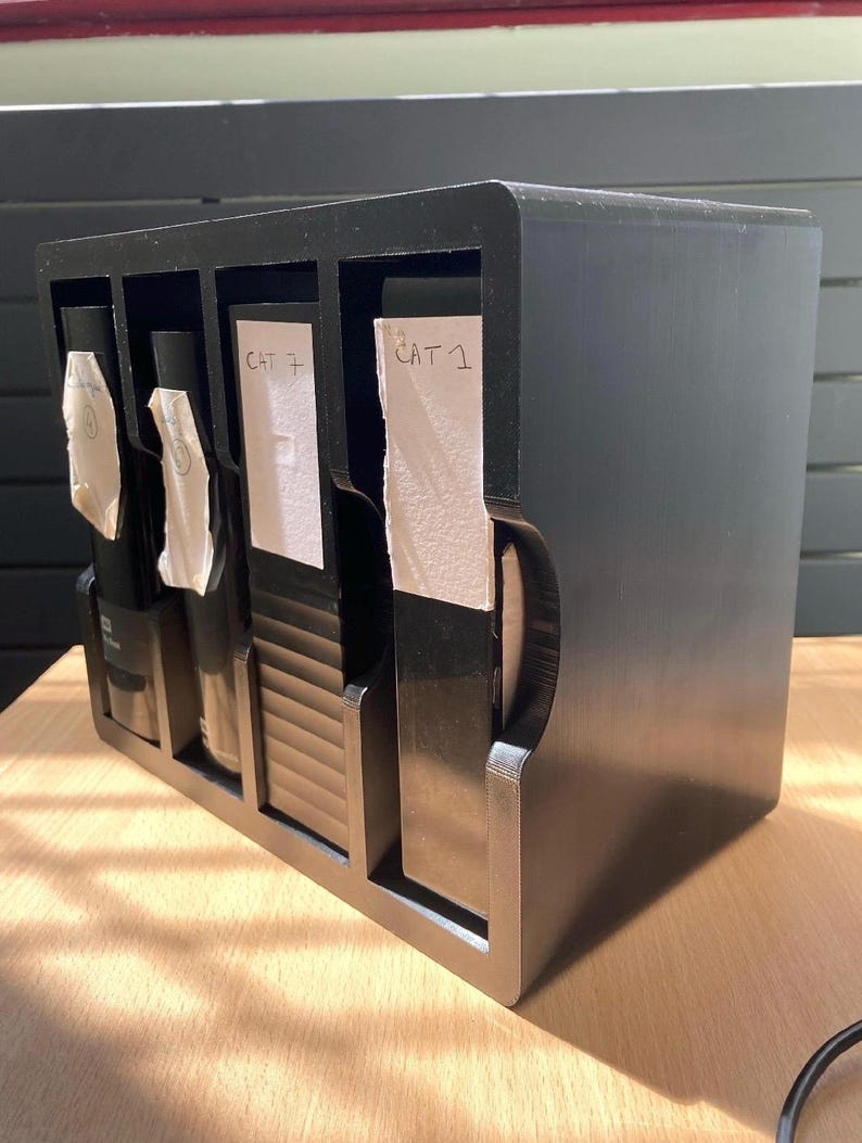 May include: A black, multi-compartment organizer with four sections. Each section holds a dark-colored bottle or item, with white labels reading "CAT 7" and "CAT 1". The organizer is made of a solid, dark material and sits on a light-colored wooden surface.