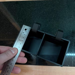 Puede incluir: Un contenedor de almacenamiento negro y rectangular con dos compartimentos y dos clips. El contenedor mide aproximadamente 10 cm de ancho. La imagen est&aacute; tomada desde arriba, mostrando el contenedor sobre una superficie de madera y negra.