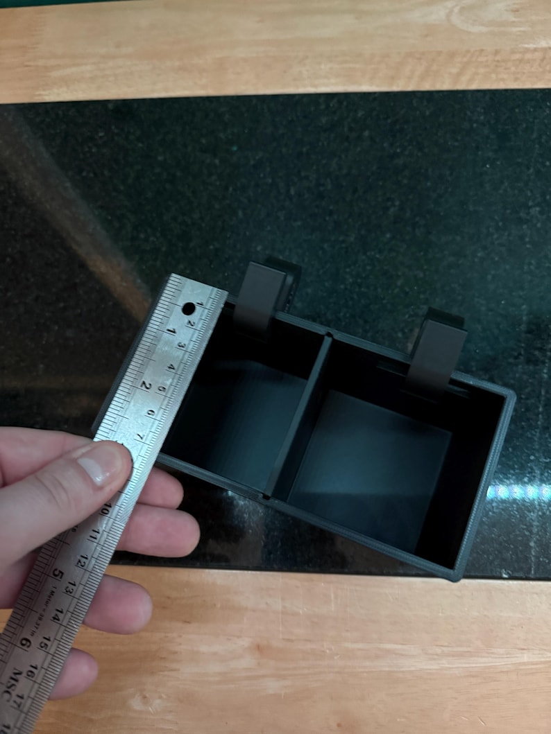 Puede incluir: Un contenedor de almacenamiento negro y rectangular con dos compartimentos y dos clips. El contenedor mide aproximadamente 10 cm de ancho. La imagen est&aacute; tomada desde arriba, mostrando el contenedor sobre una superficie de madera y negra.