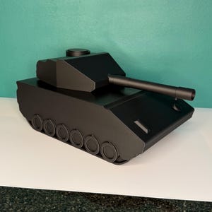 May include: A matte black toy tank featuring a rotating turret, a long barrel, and a series of circular wheels along the side. The tank has a geometric, angular design with a flat top and a smooth, solid black finish across the entire structure.