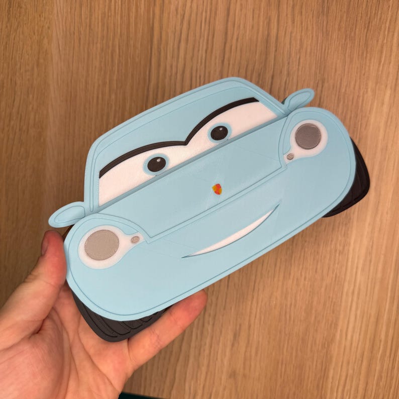 May include: A light blue car-shaped coaster with a smiling face, black tires, and white accents. The coaster has a cartoon design and is held in a hand.