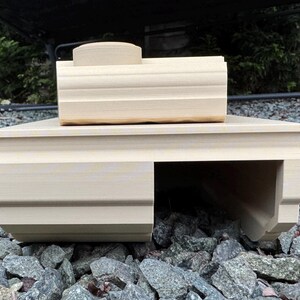 May include: A light beige, 3D-printed plastic tank model featuring a rectangular base with a hollow tunnel section on the right side and a smaller, elevated rectangular turret component positioned on top of the main body structure.