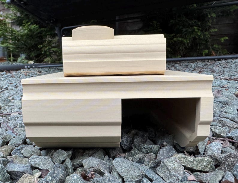 May include: A light beige, 3D-printed plastic tank model featuring a rectangular base with a hollow tunnel section on the right side and a smaller, elevated rectangular turret component positioned on top of the main body structure.