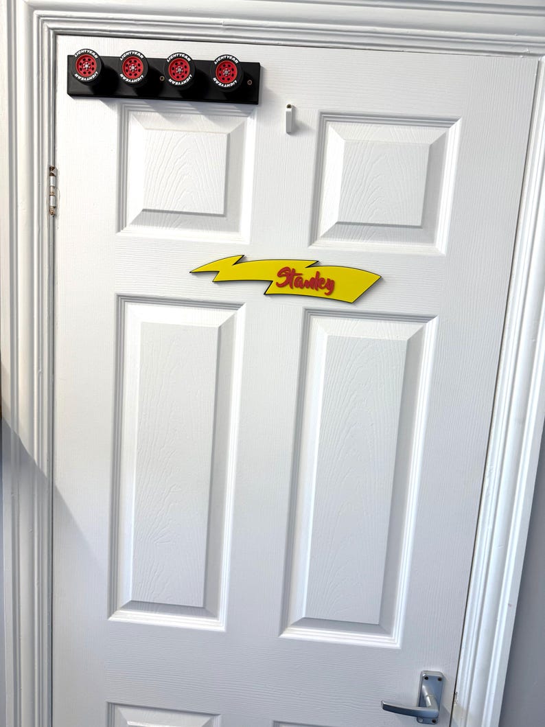 May include: A white door with a black wall-mounted rack holding four red and black decorative lights. Below, a yellow arrow-shaped sign with the name "Stanley" in red lettering.