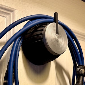 Op de afbeelding: Een 3D-geprinte kabelorganisator in de vorm van een knop, met een zwarte en zilveren afwerking, gemonteerd op een witte deur. Blauwe kabels zijn om de organisator gewikkeld.