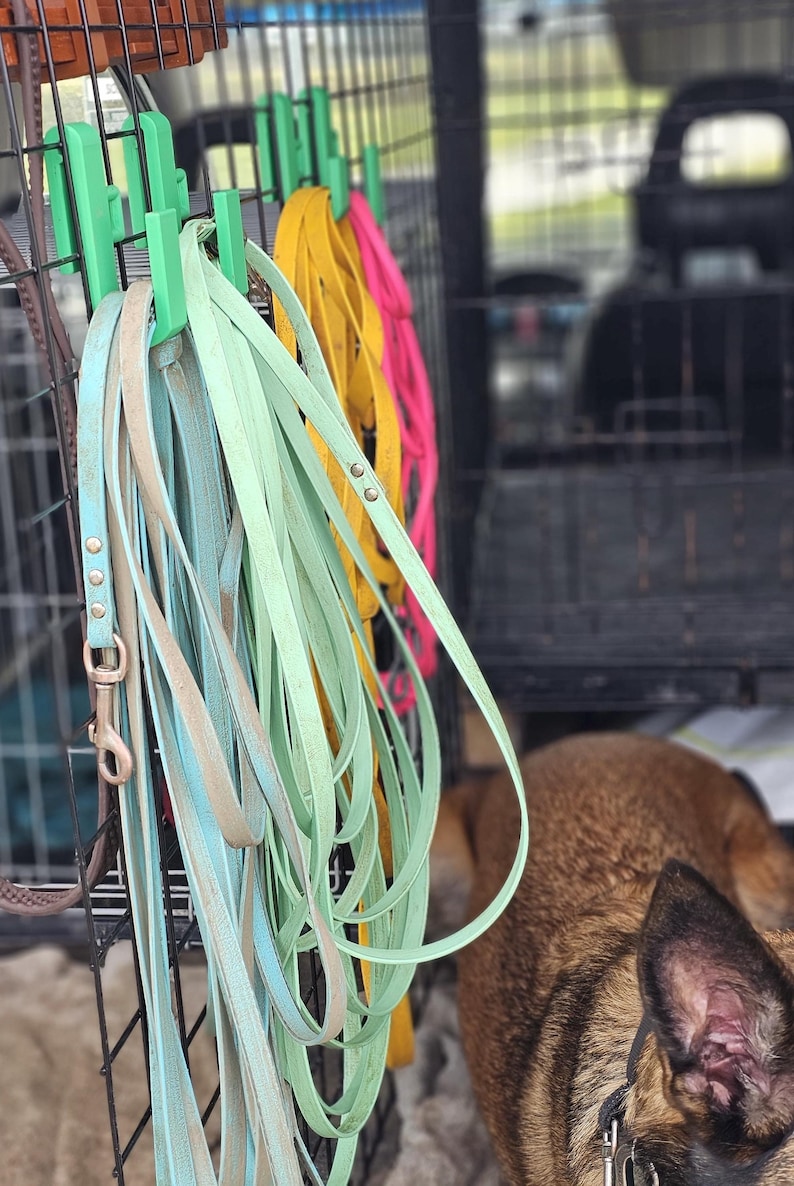 Green 3D-printed hooks attached to a dog crate, holding multiple colourful longlines in blue, yellow, and pink. A Belgian Malinois dog is partially visible in the foreground inside the crate.