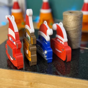 Może przedstawiać: Kolekcja kolorowych ozd&oacute;b choinkowych w kształcie samochod&oacute;w, każda ozdobiona czapką Świętego Mikołaja. Ozdoby są czerwone, brązowe i niebieskie, z białymi i czerwonymi czapkami Świętego Mikołaja. W tle szpula sznurka.